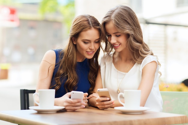 technology, lifestyle, friendship and people concept - happy young women or teenage girls with smartphones and coffee cups at cafe outdoors