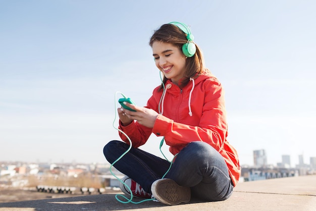 Concetto di tecnologia, stile di vita e persone - giovane donna sorridente o adolescente con smartphone e cuffie che ascolta musica all'aperto