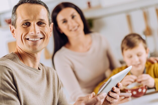 Foto amante della tecnologia. l'attenzione si concentra su un giovane affascinante che utilizza un tablet e posa per la fotocamera mentre la sua famiglia si lega durante la colazione in sottofondo