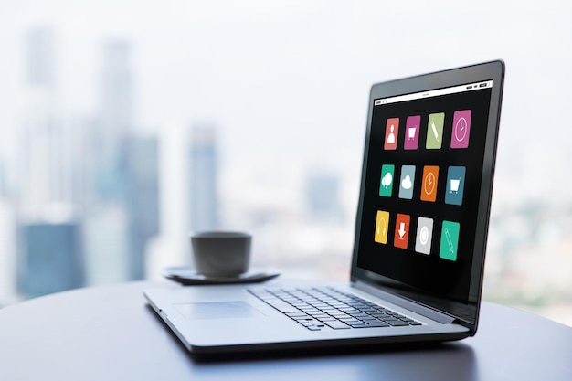 Photo technology, media, business and modern life concept- close up of open laptop with computer menu icons on screen and coffee cup on table at office or hotel room