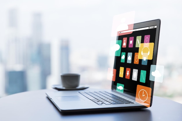 technology, media, business and modern life concept- close up of open laptop with computer menu icons on screen and coffee cup on table at office or hotel room