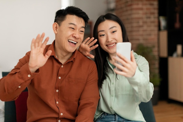 Technology meeting remotely Happy asian couple having video call and waving hands at cellphone webcamera