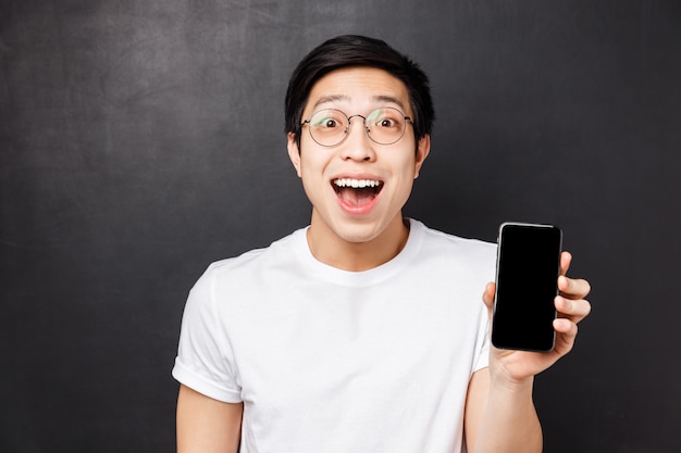 Technology, messaging and people concept. Close-up portrait of happy, surprised and impressed young excited asian man in white t-shirt, showing smartphone display amazed with cool app