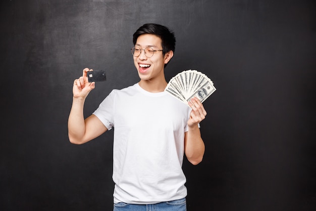 Technology, money and prizes concept. Portrait of boastful smiling, happy asian guy become rich, lucky to win prize, holding dollars and credit card, making decison how invest money, 