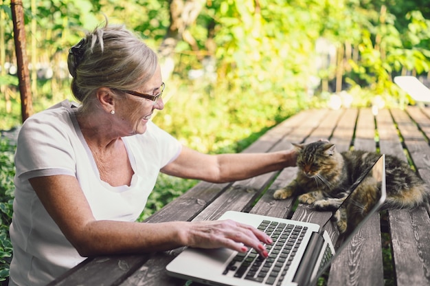 Technology, old age people concept - elderly happy senior woman with home cat working online with laptop computer outdoor in the garden. Remote work, distance education.