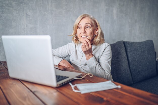 Technology, old age and people concept - happy older senior woman with face medical mask working and making a video call with laptop computer at home during coronavirus COVID19 pandemic. Stay home