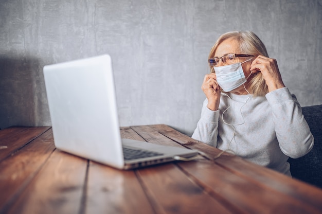 Tecnologia, vecchiaia e concetto della gente - donna senior più anziana triste sola nella maschera medica del fronte che lavora e che fa una video chiamata con il computer portatile a casa durante la pandemia di covid19 del coronavirus. stare a casa