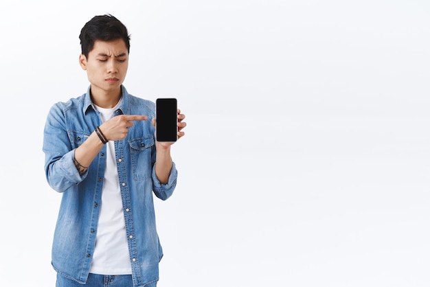 기술 온라인 라이프 스타일 개념 의심스럽고 의심스러운 젊은 아시아 남성의 초상화가 곁눈질로 스마트폰을 가리키며 일부 불신이 있는 휴대 전화 디스플레이를 보고 있습니다.