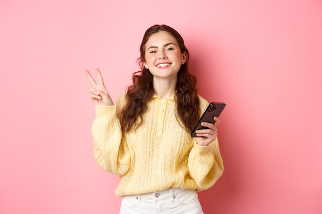 Technology and online shopping. Happy young woman smiling pleased, holding smartphone, showing v-sign and look positive at camera, standing over pink wall.