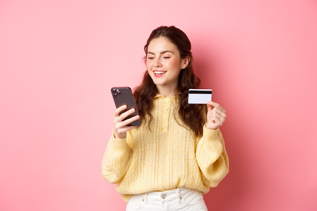 Technology and online shopping. young pretty lady paying online with credit card, looking at smartphone and smiling, standing over pink wall.