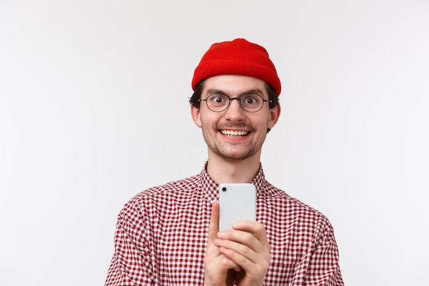 Technology and people concept. Excited handsome young hipster guy attend awesome concert and record it on mobile phone, holding phone smiling amused, stand white wall