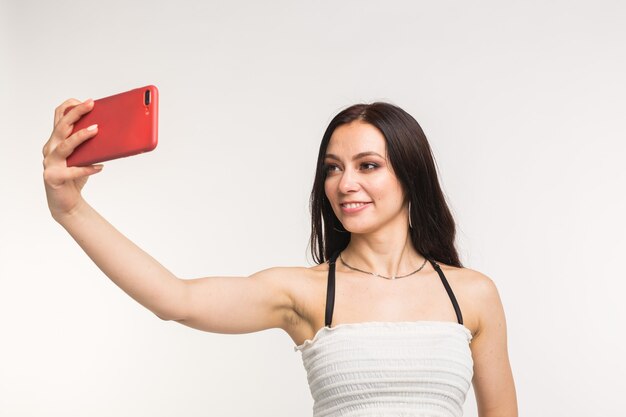 Technology and people concept - young woman making a selfie photo and smiling on white wall.