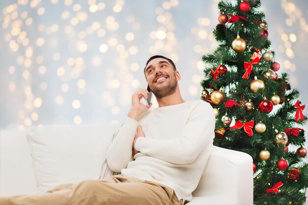 technology, people and holidays concept - smiling man calling on smartphone over christmas tree and lights background