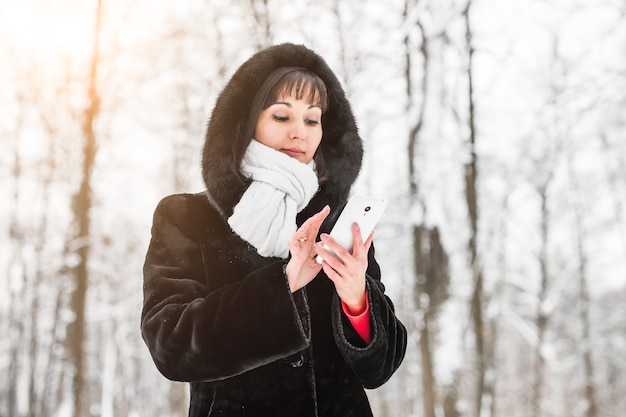 Technology, people and winter concept - Young woman with smartphone and winter landscape snowflakes