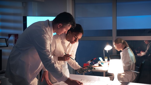 Technology research laboratory mixed race electronics engineers in white coats working with electronic board and control electronics scheme in lab