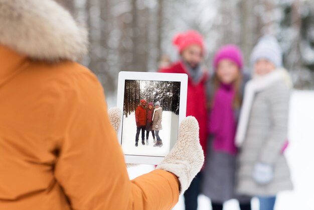 Technology, season, friendship and people concept - close up of happy men and women taking picture with tablet pc computer in winter forest