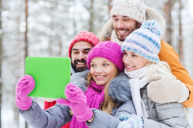 technology, season, friendship and people concept - group of smiling men and women taking selfie tablet pc computer in winter forest