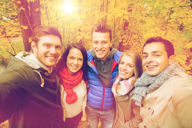 テクノロジー、季節、友情、人々のコンセプト-秋の公園でスマートフォンやカメラで自分撮りをしている笑顔の男性と女性のグループ