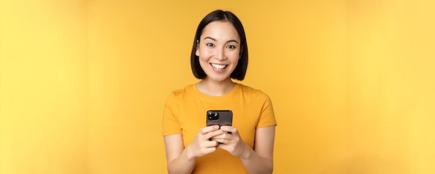 Photo technology smiling asian woman using mobile phone holding smartphone in hands standing in tshirt against yellow background