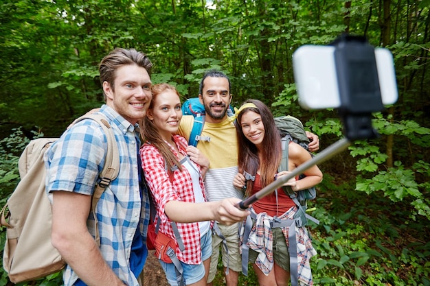 technology, travel, tourism, hike and people concept - group of smiling friends walking with backpacks taking picture by smartphone on selfie stick in woods