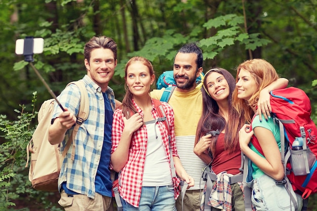 technology, travel, tourism, hike and people concept - group of smiling friends walking with backpacks taking picture by smartphone on selfie stick in woods