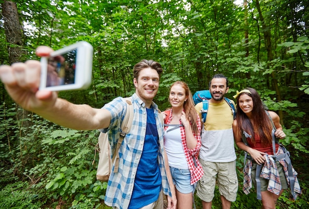 技術、旅行、観光、ハイキング、人々 のコンセプト - 森の中でスマート フォンで selfie を取ってバックパックを持って歩く笑顔の友人のグループ