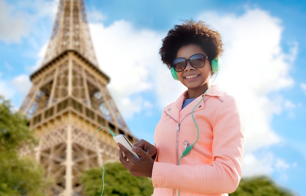 テクノロジー、旅行、観光、人のコンセプト – エッフェル塔の背景にスマートフォンとヘッドフォンで音楽を聴く笑顔のアフリカ系アメリカ人の若い女性または10代の女の子