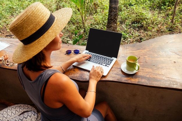 テクノロジーと旅行屋外での作業フリーランスのコンセプト熱帯のビーチのカフェでラップトップを使用して帽子をかぶったかなり若い女性