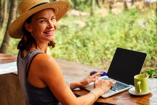 Technology and travel Working outdoors Freelance concept Pretty young woman in hat using laptop in cafe on tropical beachHooray victory success and successful deal concept promotion at work