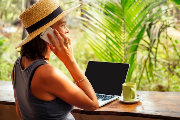 Technology and travel Working outdoors Freelance concept Pretty young woman in hat using laptop in cafe on tropical beachHooray victory success and successful deal concept promotion at work