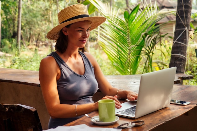 기술 및 여행 야외에서 작업 프리랜서 개념 열대 해변에 있는 카페에서 노트북을 사용하는 모자를 쓴 예쁜 젊은 여성Horay 승리 성공 및 직장에서의 성공적인 거래 개념 프로모션