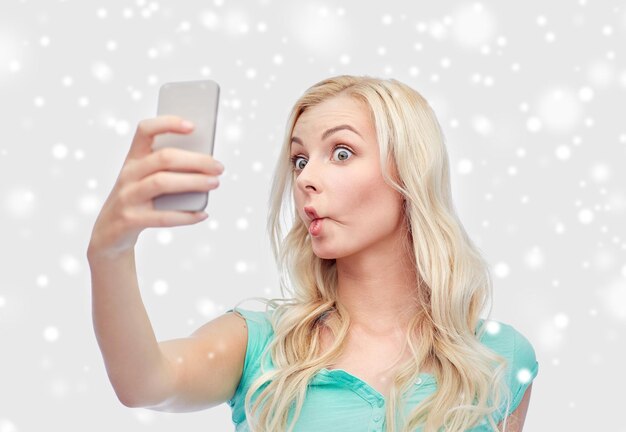 Premium Photo  Technology, winter holidays, christmas, fun and people  concept - funny young woman or teenage girl taking selfie with smartphone  and making fish face over snow