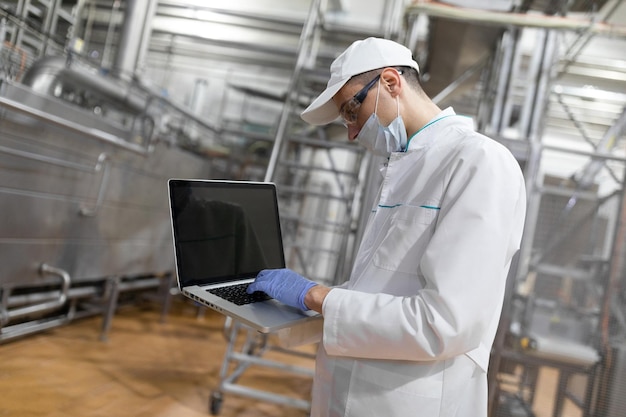 Technoloog in een witte jas masker en handschoenen met een laptop in zijn handen is in de winkel voor de productie van boter en kaas productieproces in de zuivelfabriek