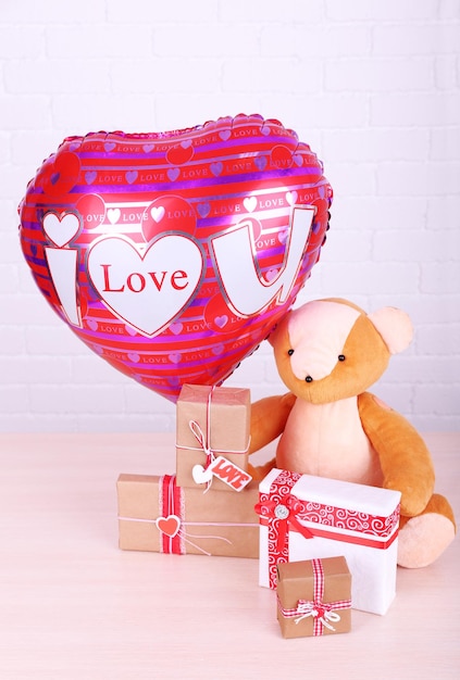 Teddy bear takes gifts and love heart balloon on wooden table, on the brick wall background