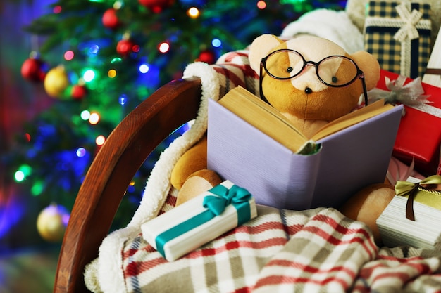 Плюшевый мишка с книгой и подарочными коробками в кресле-качалке на фоне елки