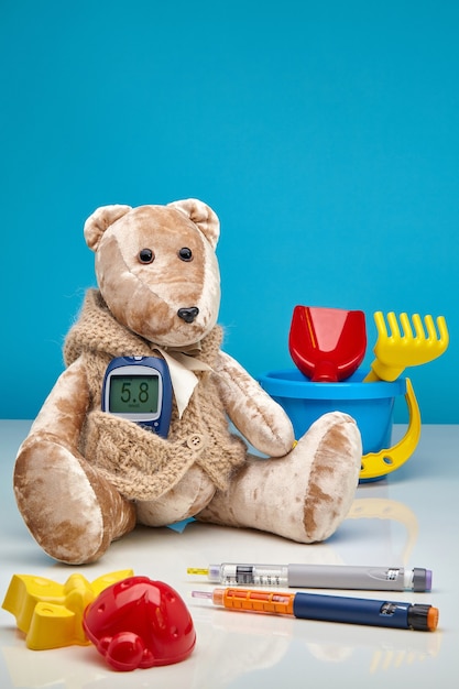 Teddy bear with a glucometer and scattered children's toys and insulin pens on a blue and white