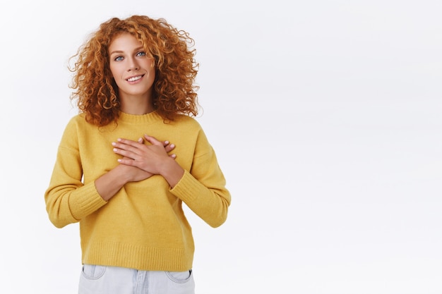 Tedere, aangeraakte roodharige blanke vrouw met krullend gemberhaar, druk op de armen op het hart en zuchtend glimlachend dankbaar, kijk blij en gevleid als romantisch cadeau, witte muur