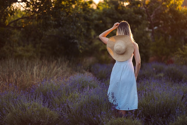 Tedere vrouw staat met haar rug in witte jurk en hoed tussen lavendel en verlicht door zonnestralen