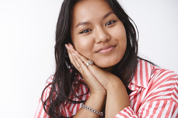 Tederheid, schoonheid en lichaam positief concept. portret van een lieftallige en zachtaardige vriendin die met haar hoofd op de handpalmen leunt, dromerig en schattig naar de camera staart, tevreden met een tedere blik over de witte muur