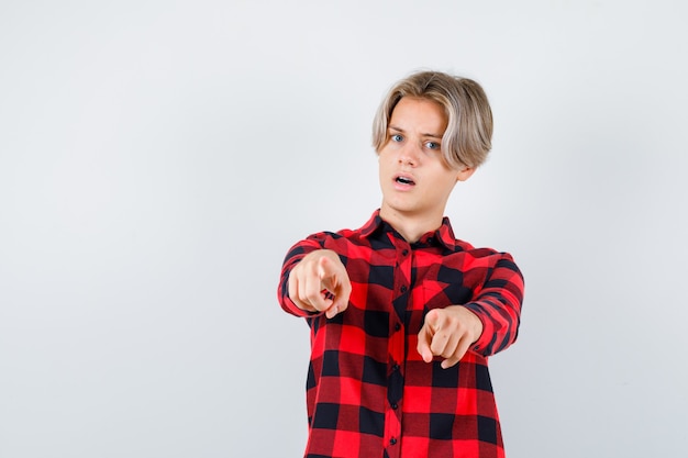 Maschio biondo teenager in camicia casuale che indica davanti e che sembra perplesso, vista frontale.