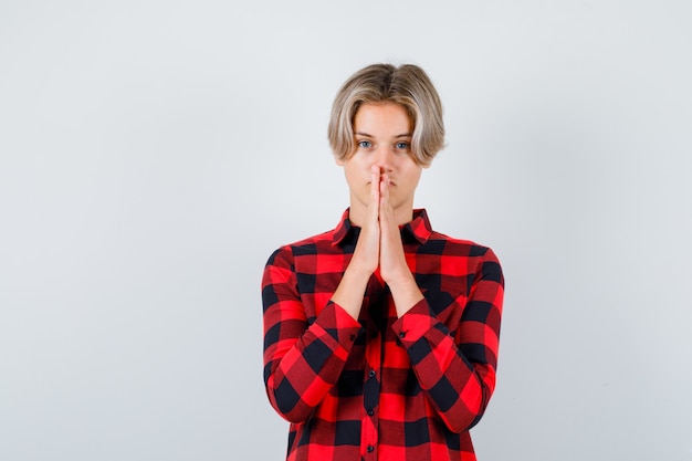 Teen blond male with hands in praying gesture in casual shirt and looking hopeful , front view.