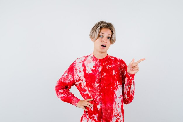 Teen blonde male in oversized shirt pointing right, with hand on waist and looking thoughtful , front view.