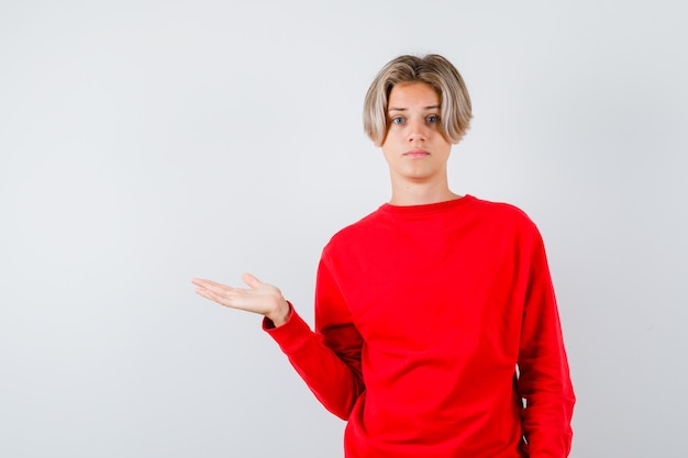 Photo teen blonde male spreading palm aside in red sweater and looking thoughtful , front view.