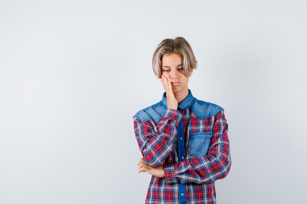 Teen boy in checkered shirt with hand on cheek and looking disappointed , front view.