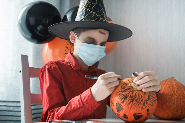 Photo teen boy in costume and face masks to protect against covid-19 drawing a pumpkin for the halloween celebration