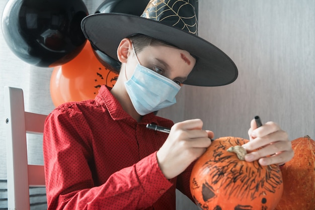 Teen boy in costume preparing for the Halloween celebration drawing a pumpkin. Halloween carnival with new reality with pandemic concept