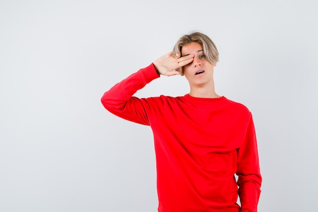 Teen boy holding fingers on his eye, opening mouth in red sweater and looking surprised. front view.