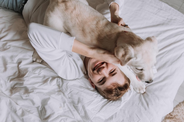 十代の少年は犬の上面図で白い寝具の上にベッドに横たわっていますペットは朝に所有者を起こします