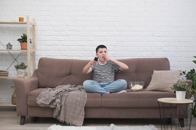 Photo a teen boy is sitting on the sofa eating pop corn holding control panel and watching tv