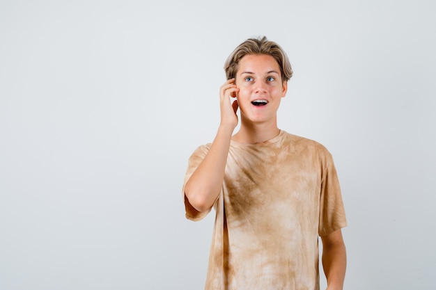 Teen boy keeping hand on head while opening mouth in t-shirt and looking amazed. front view.